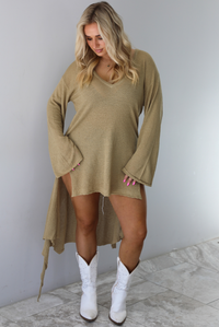 Woman wearing a camel long-sleeve top that's tied in the front with a knot and has a train in the back. The photo shows a closeup of the top which has small holes in it. The top is untied to show the full length of the front, which stops mid-thigh on the 5'8" model. The photo is taken indoors against a white wall with white marble flooring. 