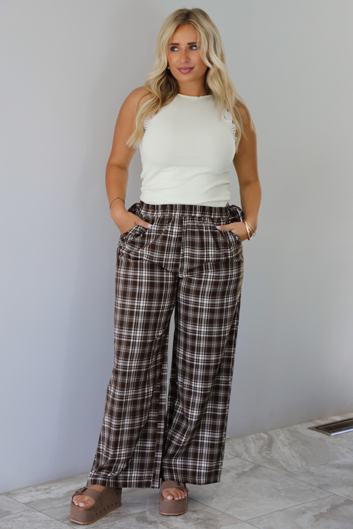 A woman modeling a pair of brown and sand plaid wide-leg pants with a cream ribbed sleeveless top, standing indoors against a white wall with white marble flooring. 