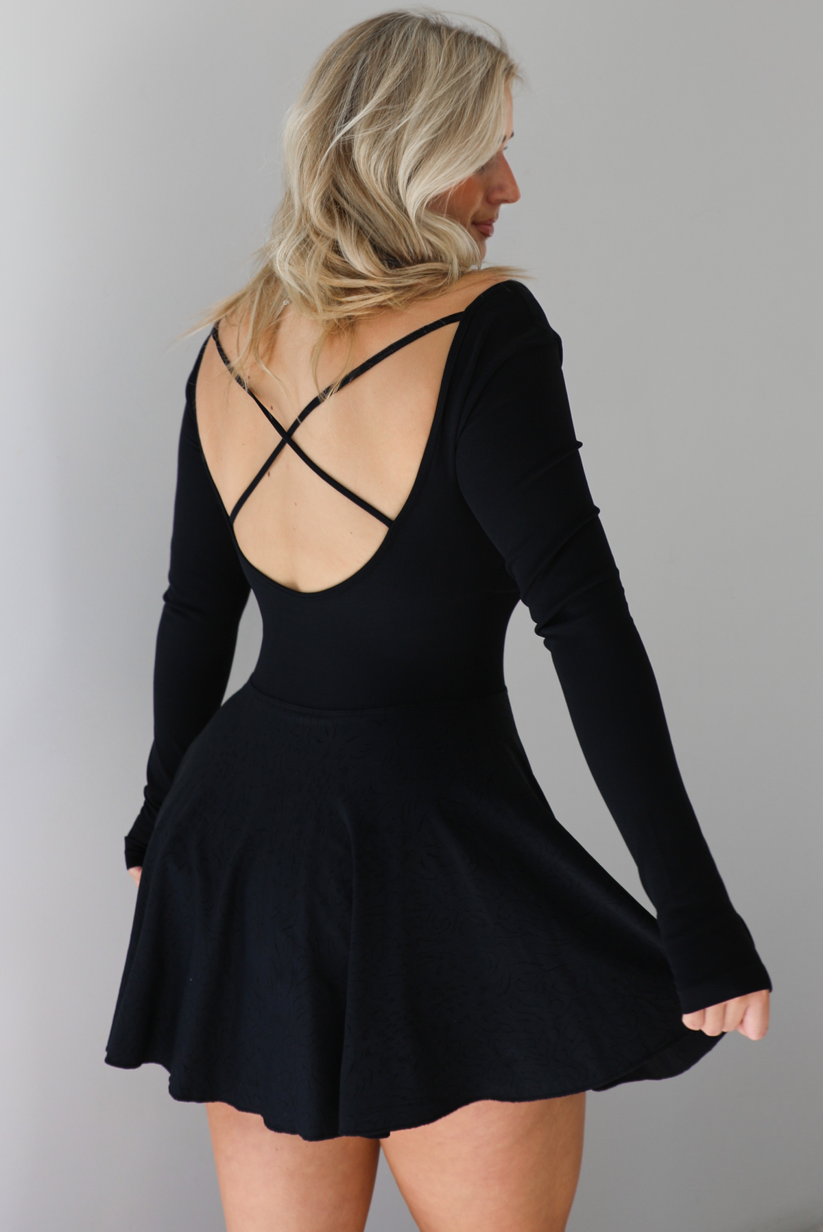 Woman wearing a black athleisure dress that has long sleeves with thumbhole cutouts, a scooped neckline, fitted bodice, and flowy skirt. The photo shows a closeup of the back of the dress, which has an open back design with a crisscross detail. The photo is taken indoors against a white wall.