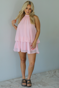 Woman wearing a pink dress that has a halter tie neckline, a cinched detail at the hips, and a flowy design indoors against a white wall with white marble flooring. 