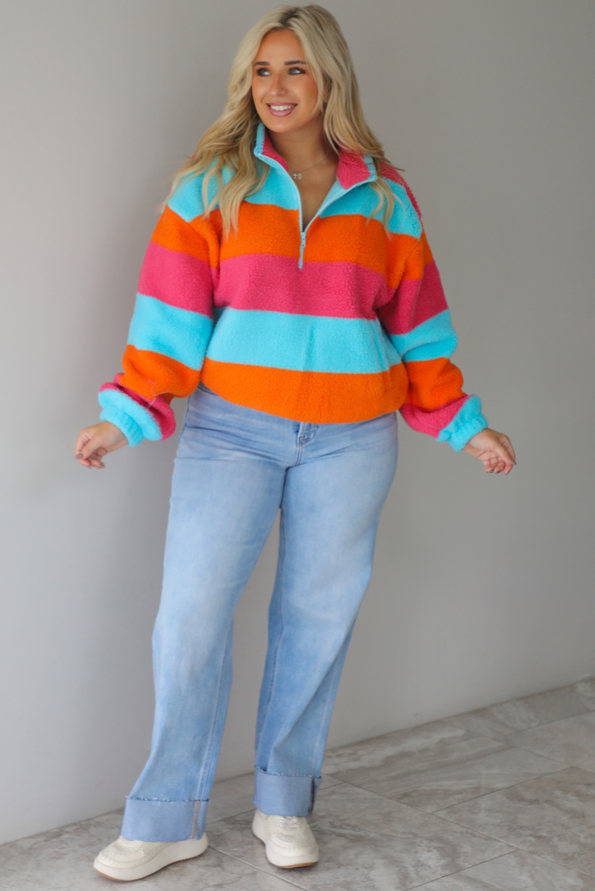 A woman wearing a turquoise, pink, and orange striped sherpa quarter-zip sweater with light wash denim jeans, standing indoors against a white wall with white marble flooring. 