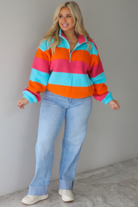 A woman wearing a turquoise, pink, and orange striped sherpa quarter-zip sweater with light wash denim jeans, standing indoors against a white wall with white marble flooring. 