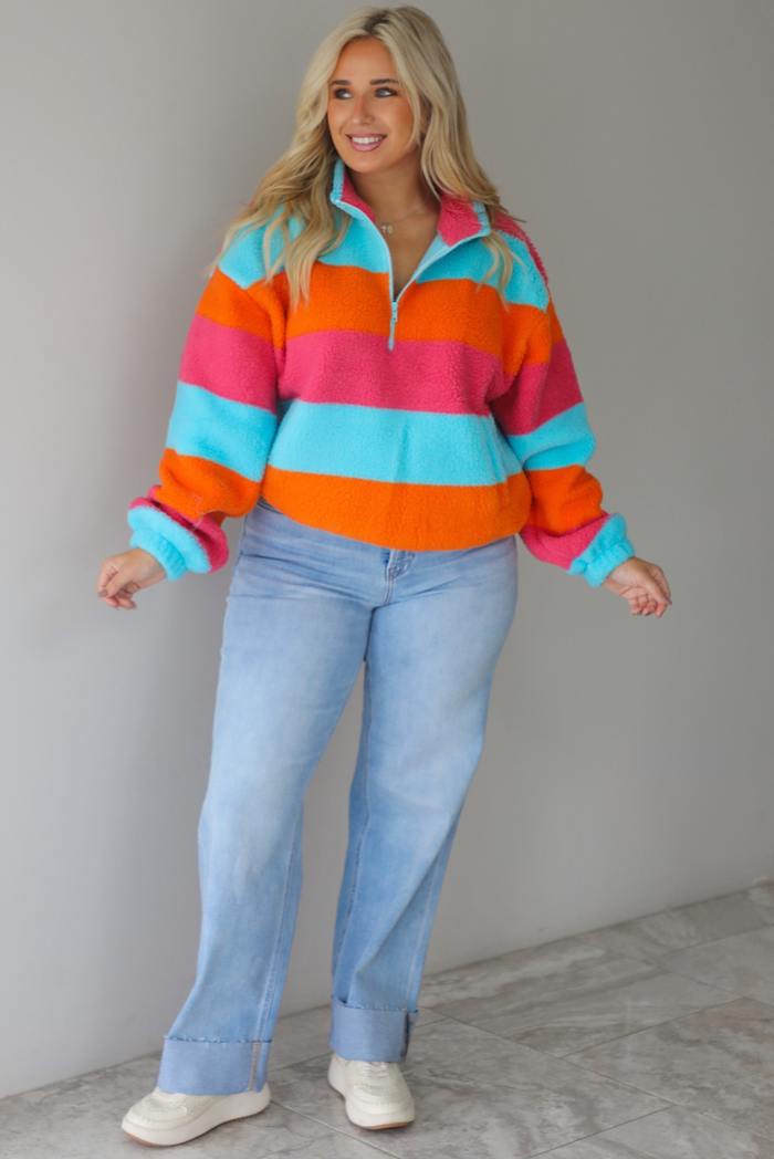 A woman wearing a turquoise, pink, and orange striped sherpa quarter-zip sweater with light wash denim jeans, standing indoors against a white wall with white marble flooring. 