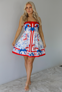 Woman wearing a white dress with red and blue sea themed graphics throughout with thick straps and functional pockets on either side. The photo is taken indoors against a white wall with white marble flooring. 