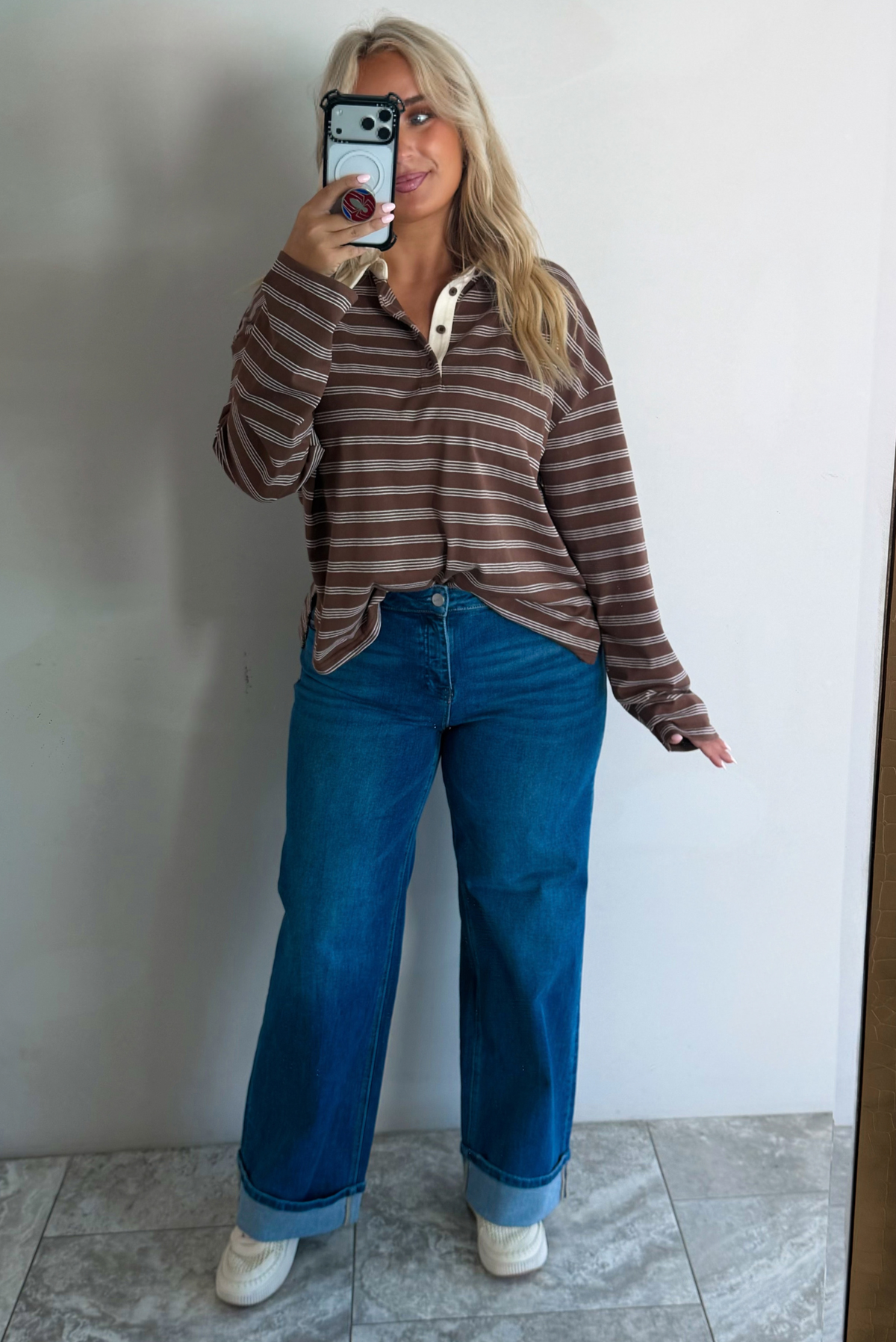 A woman wearing a brown and cream striped collared quarter-button long sleeved top, tucked into dark wash wide leg denim jeans and cream sneakers, standing in front of a white wall with white marble flooring, taking a mirror selfie. 