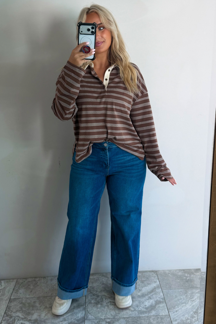 A woman wearing a brown and cream striped collared quarter-button long sleeved top, tucked into dark wash wide leg denim jeans and cream sneakers, standing in front of a white wall with white marble flooring, taking a mirror selfie. 