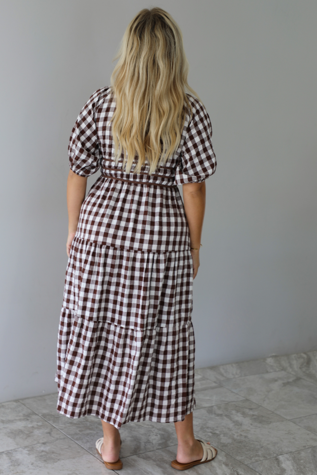 A woman shown from behind modeling a brown and white gingham tiered midi dress with puff sleeves and a smocked waist, paired with beige slide sandals, standing indoors against a white wall with white marble flooring. 