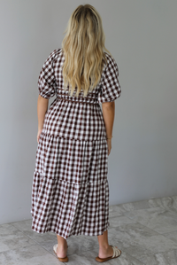 A woman shown from behind modeling a brown and white gingham tiered midi dress with puff sleeves and a smocked waist, paired with beige slide sandals, standing indoors against a white wall with white marble flooring. 