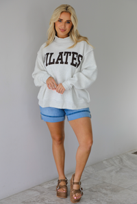 Woman wearing a heather grey oversized sweatshirt with the word 'PILATES' written in collegiate font on the front with medium wash denim shorts indoors against a white wall with white marble flooring. 