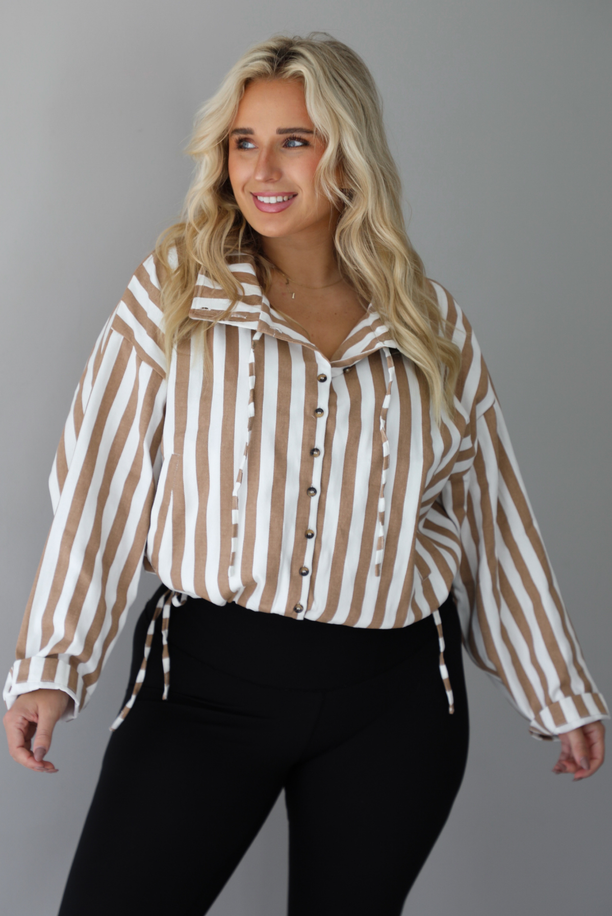 A woman wearing a tan and white striped collared long sleeved top with functional button closures along the front, worn with black leggings, standing indoors against a white wall. The top is cinched at the waist to give it a slightly cropped look. 