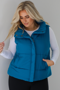 Woman wearing a teal puffer vest, white long-sleeve shirt, and black leggings indoors against a white wall. The puffer vest has a collared design, functional button closures at the neckline, a zipper closure in the front, and functional pockets on either side.