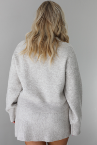 Woman wearing a grey two piece sweater set featuring a high neck sweater and a skirt. The set shows the back of the set. The photo is taken indoors against a white wall.