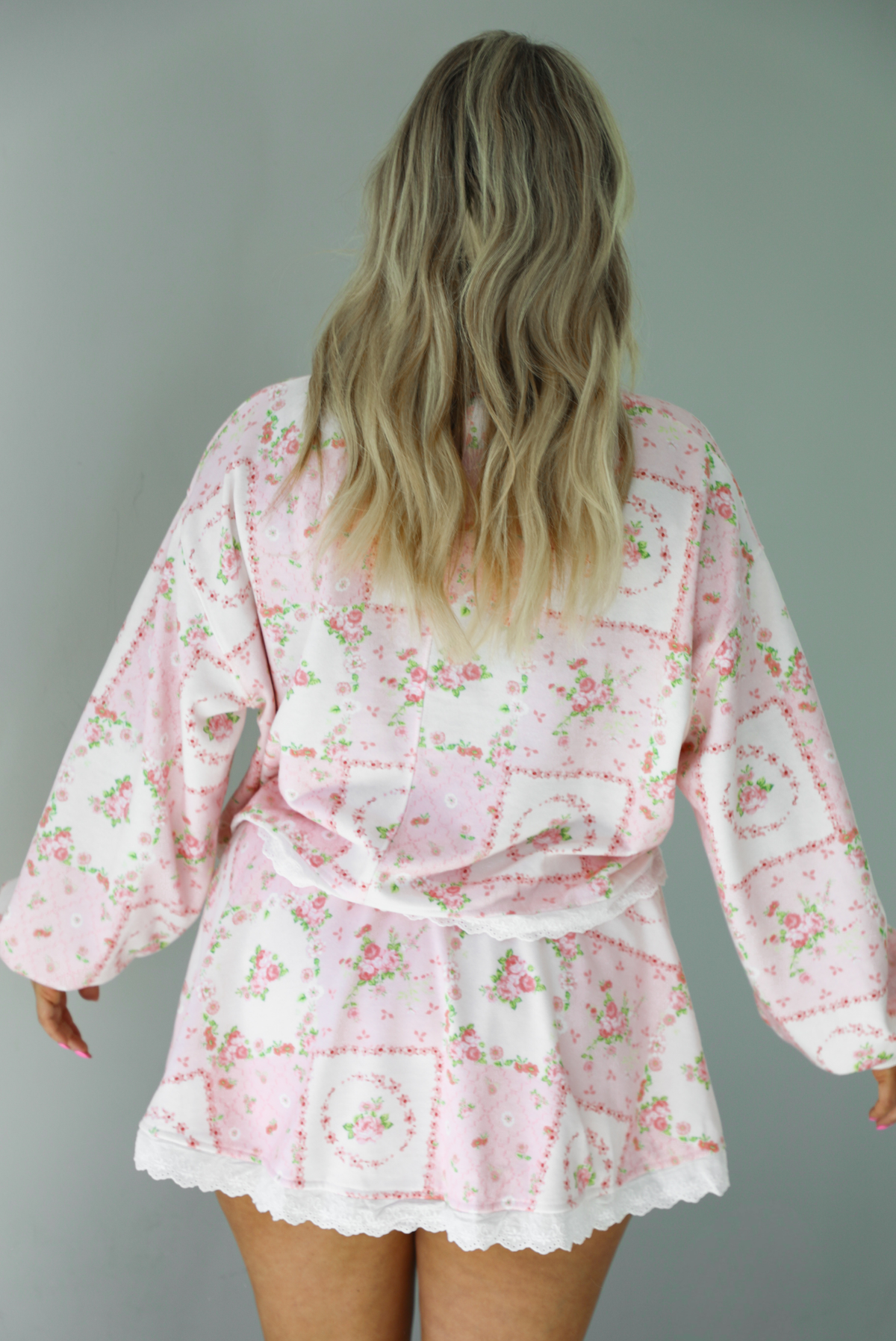 Woman wearing a two piece set featuring a pink floral set that has an oversized long sleeved top and high waisted skirt. The photo shows the back of the set. The photo is taken indoors against a white wall.  