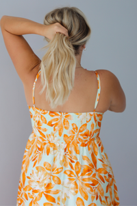 A woman shown from behind modeling a spaghetti strapped, floor-length maxi dress with a white base and a vibrant marigold orange floral pattern, standing indoors against a white wall. The photo shows the back of the dress, focusing on the smocked detail at the back and adjustable straps. 