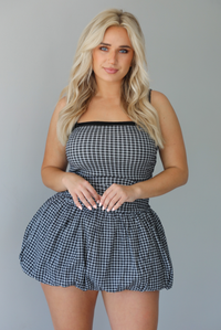 Woman wearing a black and white gingham print mini dress with a fitted, drop waist design, and a puffed hem skirt indoors against a white wall.