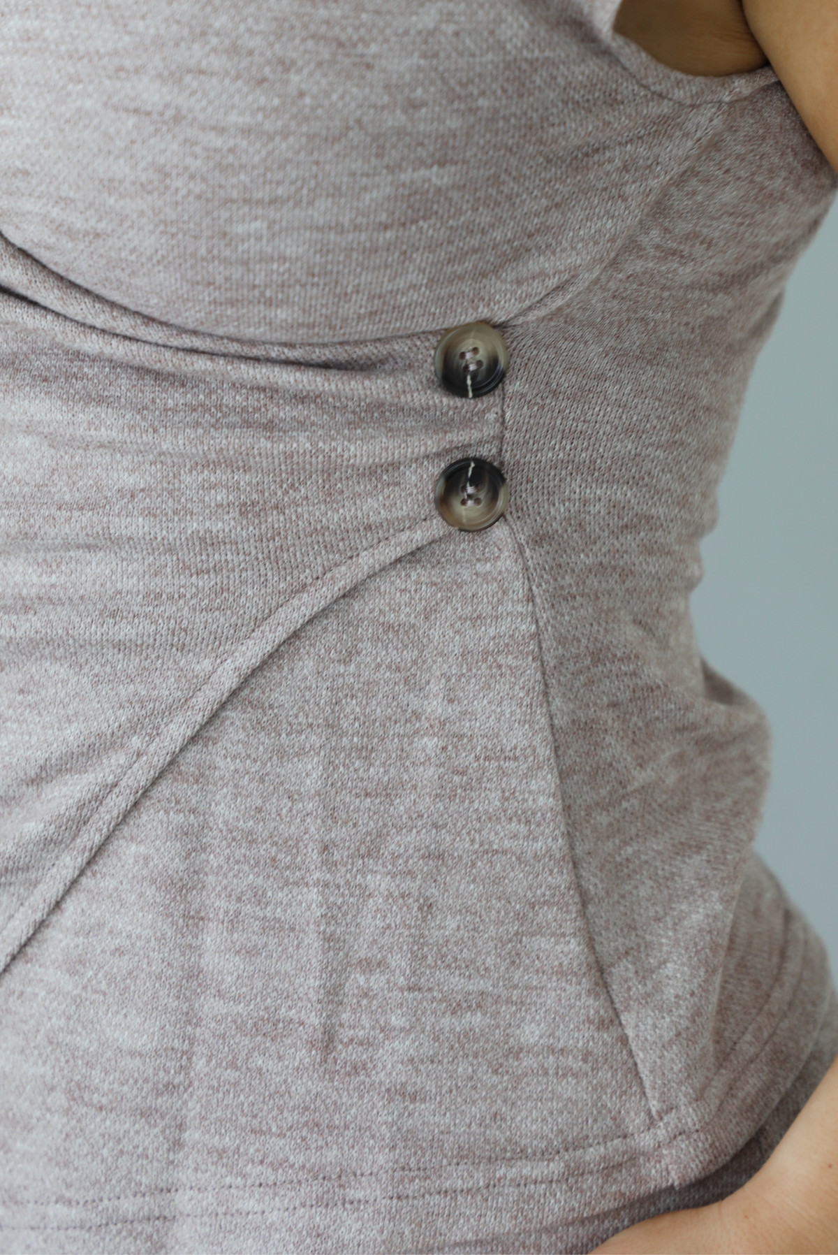 Woman wearing a taupe colored two piece set featuring a vest-appearance top with non-functional button closures on the left side and high waisted pants. The photo shows a closeup of the side of the top with the non-functional button details. The photo is taken indoors against a white wall.