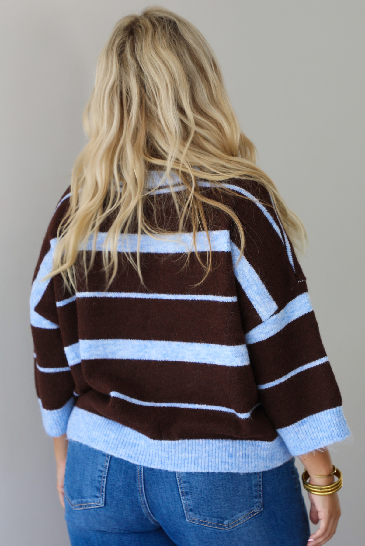 A woman shown from behind wearing a brown and blue striped knit top with dark denim jeans, standing indoors against a white wall.