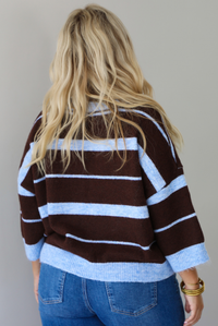 A woman shown from behind wearing a brown and blue striped knit top with dark denim jeans, standing indoors against a white wall.