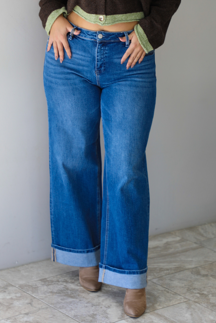 Woman wearing a pair of dark wash denim jeans, standing indoors against a white wall with white marble flooring. 