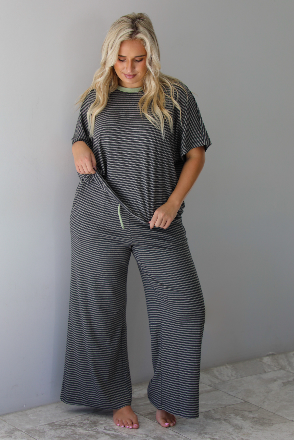 A woman wearing a charcoal and white striped two piece matching set with green trim detailing at the neckline and waistline, featuring an oversized short sleeved top and high waisted pants, standing indoors against a white wall with white marble flooring. 