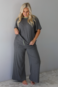 A woman wearing a charcoal and white striped two piece matching set with green trim detailing at the neckline and waistline, featuring an oversized short sleeved top and high waisted pants, standing indoors against a white wall with white marble flooring. 