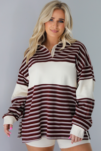 Woman wearing a burgundy and cream striped oversized collared top with cream colored biker shorts. The photo is taken indoors against a white wall.