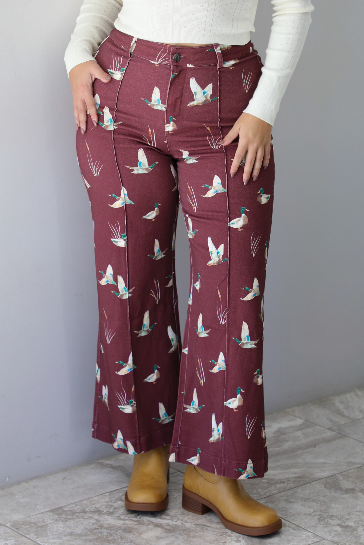 A closeup of a woman wearing a pair of maroon denim pants with a multi-colored repeating mallard design throughout have have a high waisted design, pleated details on the front, standing indoors against a white wall with white marble flooring. 