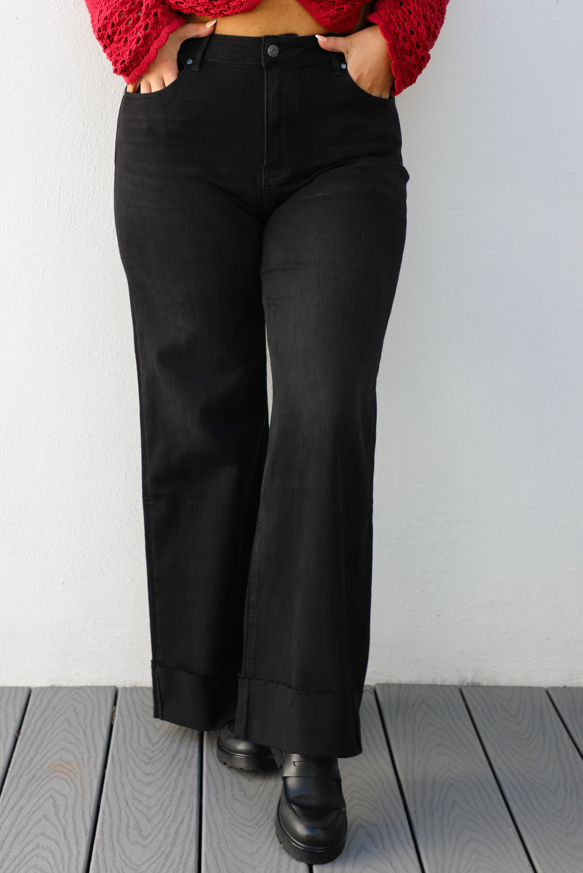 Woman model wearing a black denim jeans outdoors against a white wall. The jeans are high waisted with button and zipper closures in front, have functional front and back pockets, belt loops around, and a cuffed hem. This model is wearing a Large.