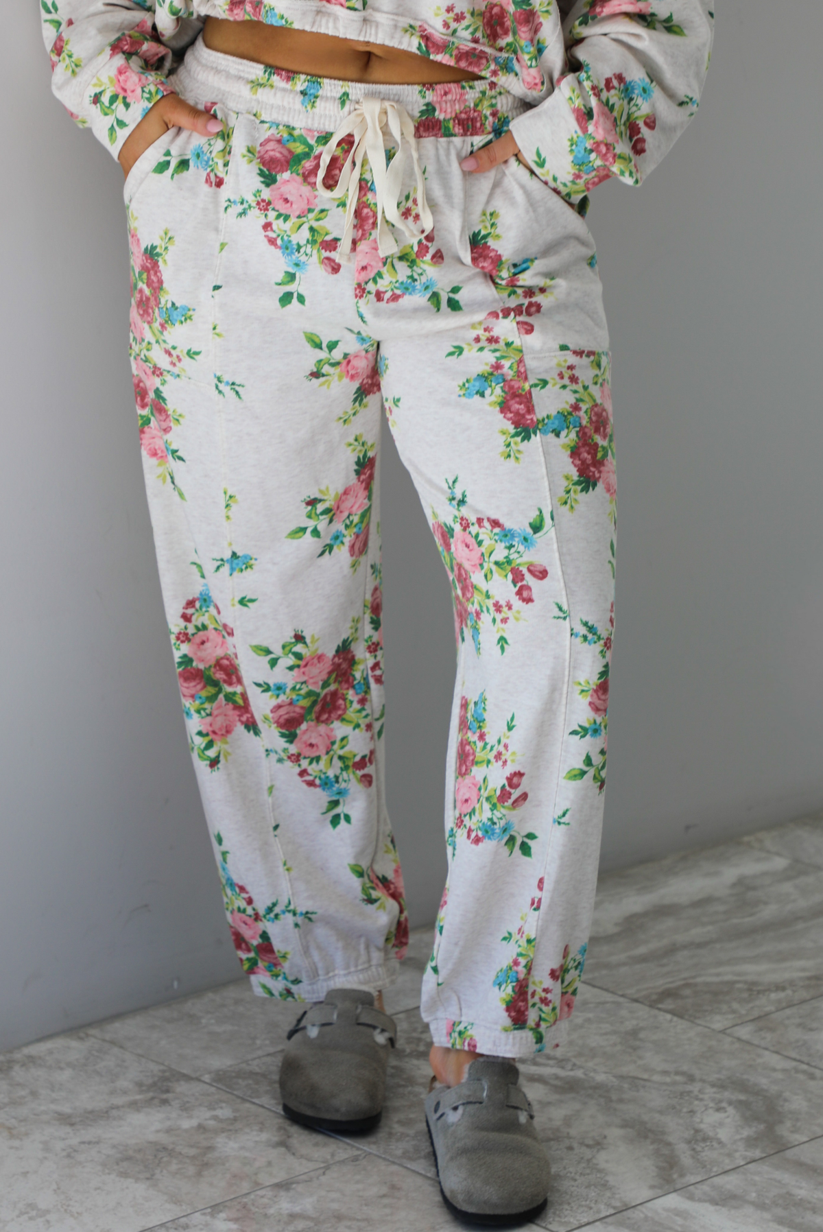 Woman wearing an oatmeal colored and multi-colored floral print two piece set featuring a collared long sleeved crop top and high waisted pants. The photo shows a closeup of the pants. The photo is taken indoors against a white wall with white marble flooring. 
