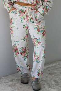Woman wearing an oatmeal colored and multi-colored floral print two piece set featuring a collared long sleeved crop top and high waisted pants. The photo shows a closeup of the pants. The photo is taken indoors against a white wall with white marble flooring. 