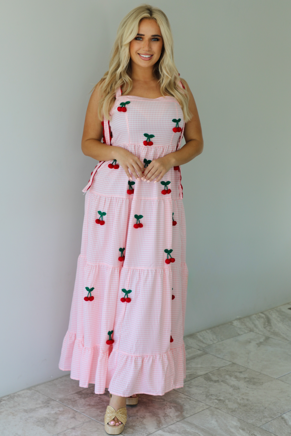 Woman wearing a pink and white gingham  maxi dress with thick straps, a fitted bodice, flowy skirt, and crocheted cherry details on the front indoors against a white wall with white marble flooring. 