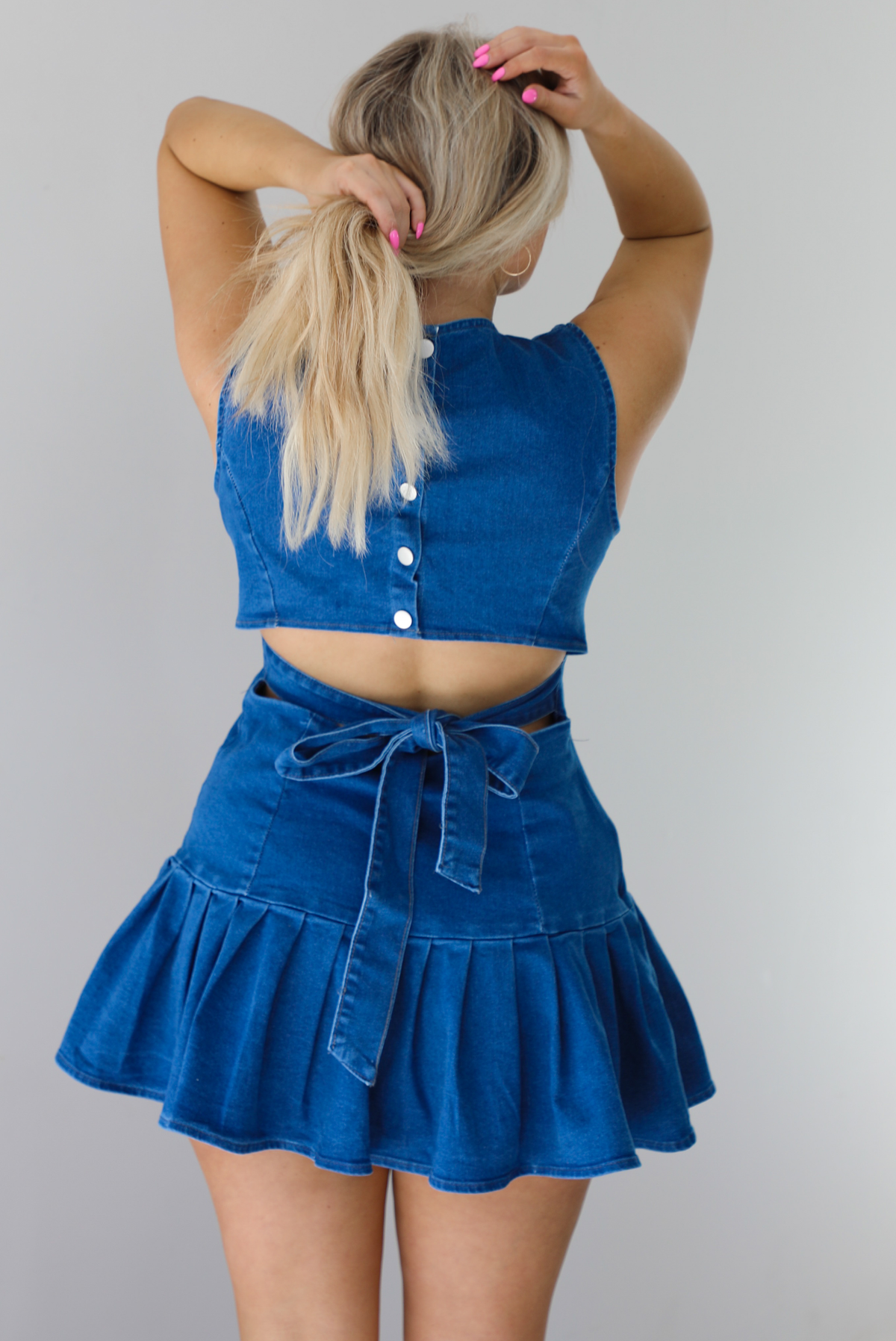 Woman wearing a dark blue denim dress that has a fitted bodice with a drop waist design thad leads to a pleated hem design. The photo shows a closeup of the back of the dress, which has button closures at the top, a cut out at the waist, and a tie at the waistline. The photo is taken indoors against a white wall.