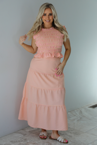 Woman wearing a pink and pale yellow gingham print two piece set featuring a smocked top and high waisted, maxi length skirt. The photo is taken indoors against a white wall with white marble flooring. 