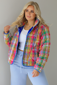 Woman wearing a multi-colored plaid, quilted style jacket paired with a white top underneath and high waisted blue lounge pants. The photo shows a closeup of the jacket, with the functional button closures in the front, functional pockets, and quilted texture. The photo is taken indoors against a white wall.