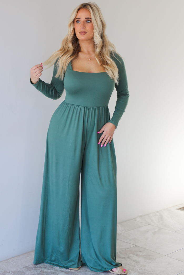 Woman wearing a green jumpsuit that has a shared neckline, long sleeved, and long, flowy pants.  The photo is taken indoors against a white wall with white marble flooring. 