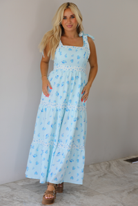 Woman wearing a light blue floral maxi dress indoors against a white wall with white marble flooring. 