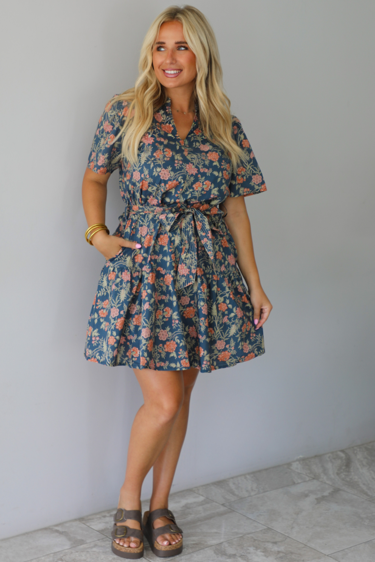 A woman modeling a short-sleeve floral mini dress with a tie waist, standing indoors against a white wall with white marble flooring. 