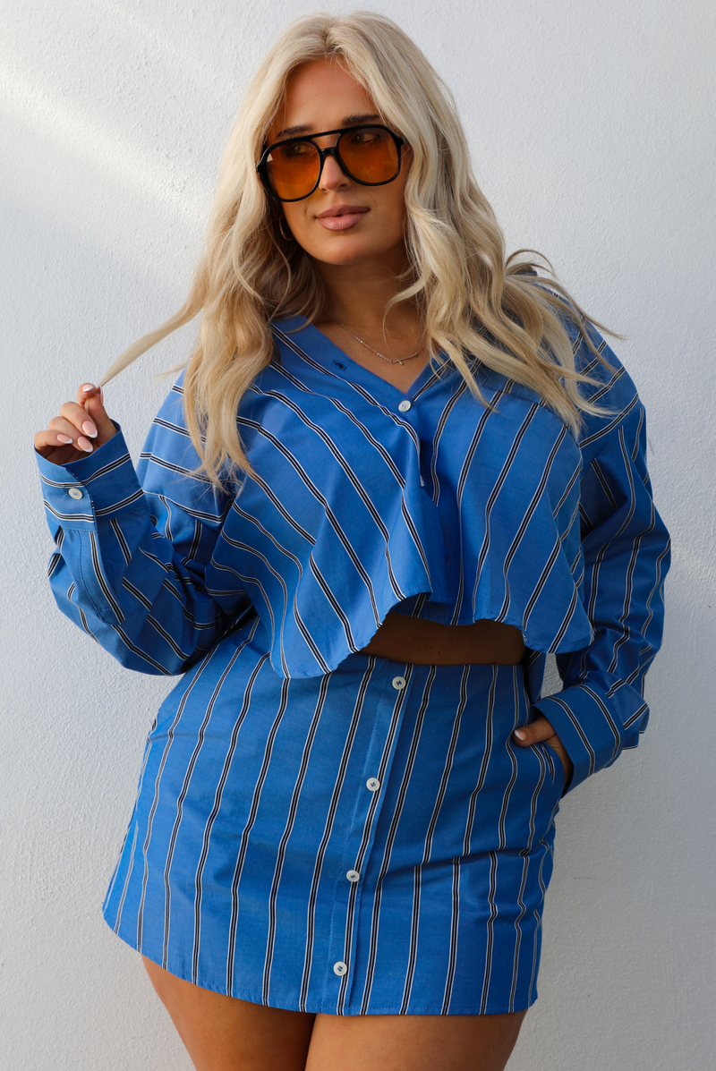 Woman wearing a blue striped set. with a white background. The set is a collared, button down cropped top with a skirt. 