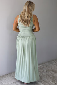 Woman wearing a sage green maxi length dress, which has a rouched detail at the bust, a fitted waistline, a flowy skirt. The photo is taken indoors against a white wall with white marble flooring and shows the back of the dress. 