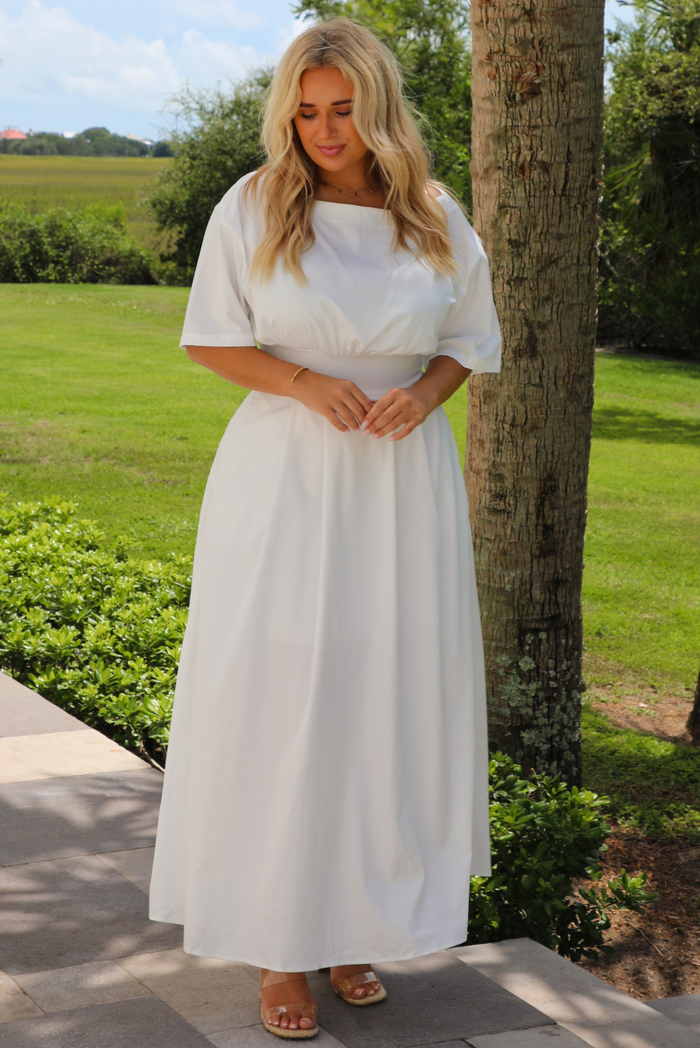 A woman wearing a white maxi dress with short puff sleeves and a cinched waist, standing outdoors on a stone path next to a tree in a grassy garden.