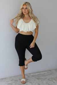 Woman wearing a cream ruffled cropped top with high waisted black, textured capri pants indoors against a white wall with white marble flooring. 