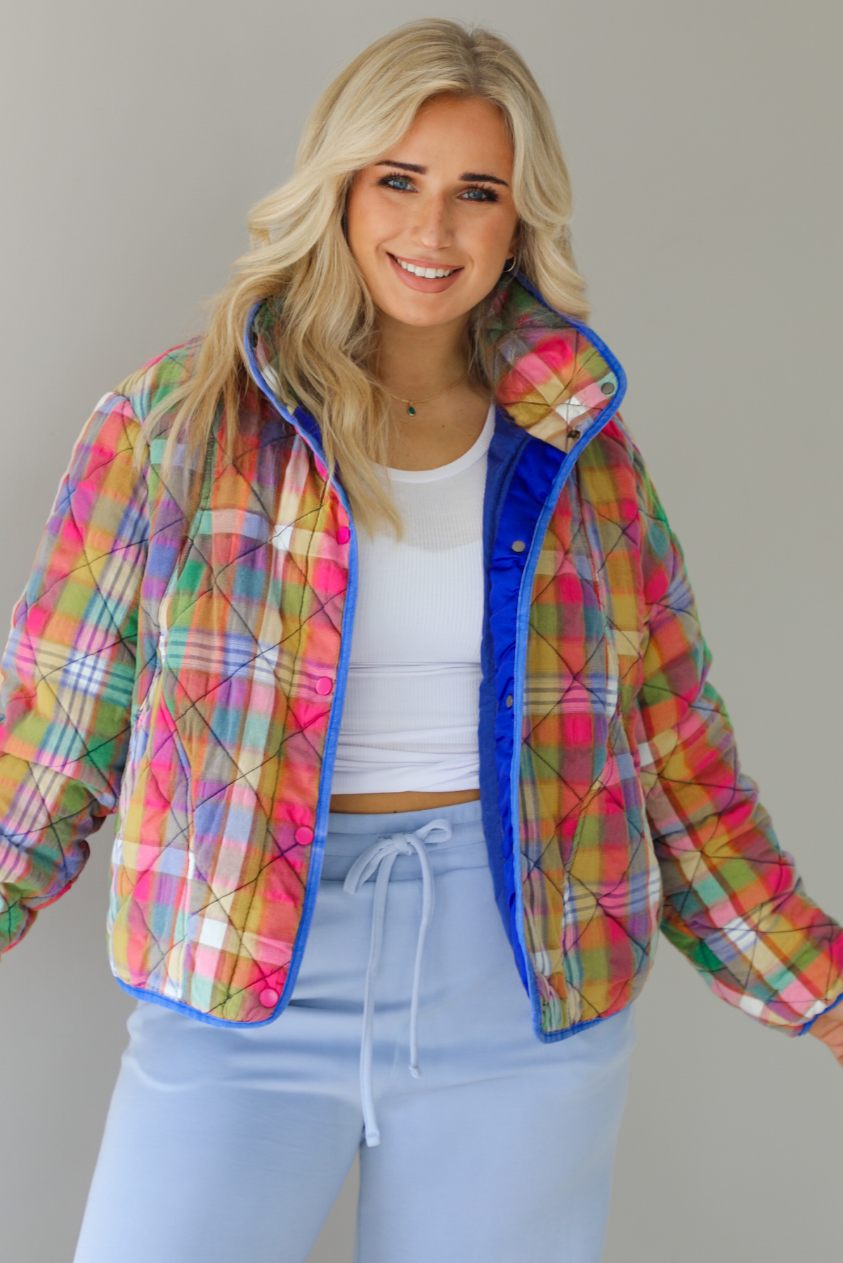 Woman wearing a multi-colored plaid, quilted style jacket paired with a white top underneath and high waisted blue lounge pants. The photo shows a closeup of the jacket, with the functional button closures in the front, functional pockets, and quilted texture. The photo is taken indoors against a white wall.