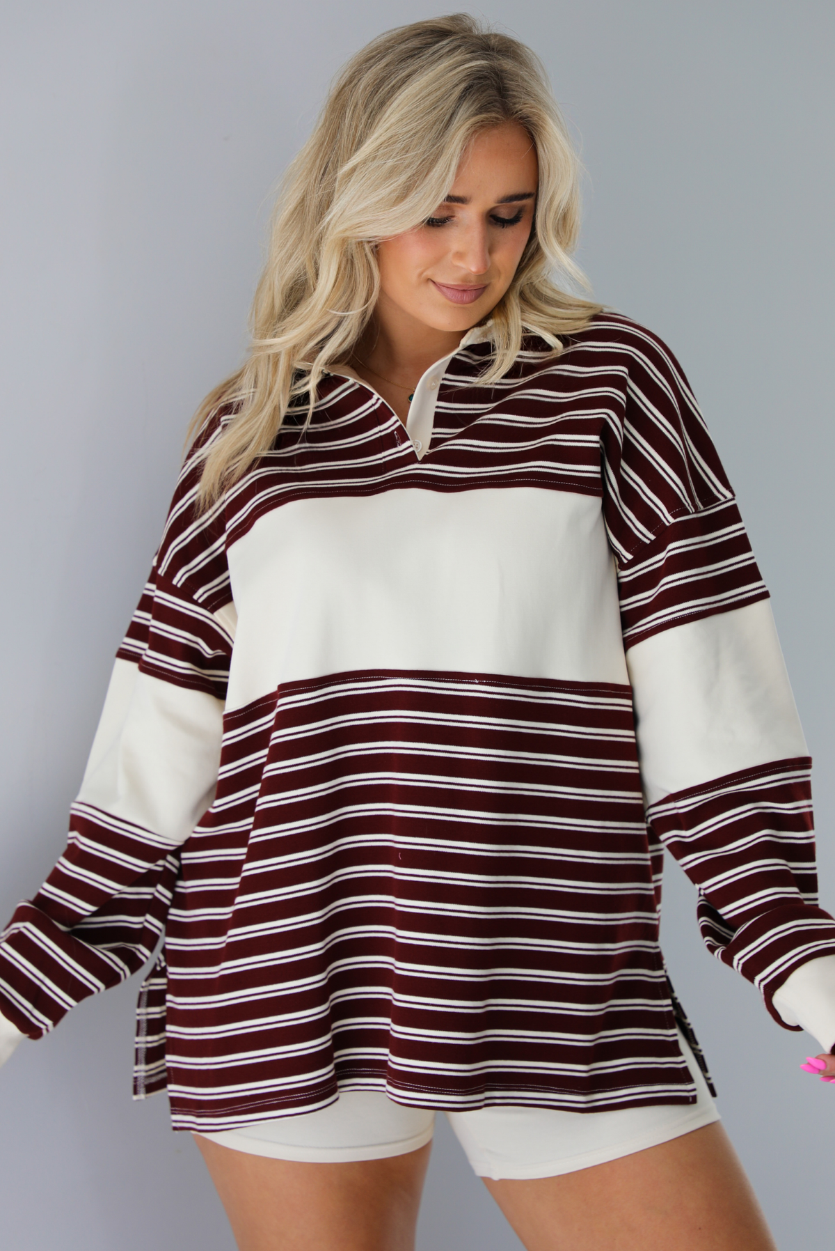 Woman wearing a burgundy and cream striped oversized collared top with cream colored biker shorts. The photo is taken indoors against a white wall.