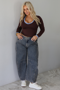 A woman wearing a dark brown, fitted, long sleeved, slightly cropped top with blue trim detailing at the v-neckline and at the sleeve hems, paired with dark acid washed barrel denim jeans, standing indoors in front of a white wall with white marble flooring.