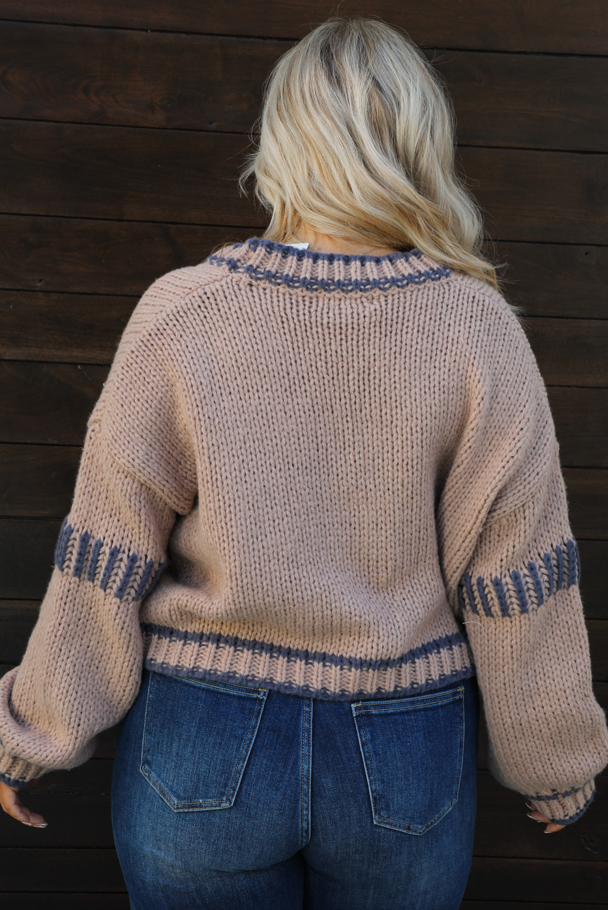 Woman model wearing a mocha brown cardigan that has grey stitching details throughout. The cardigan has a cropped cut, is oversized, and has functional button closures in the front. She's wearing the cardigan with dark denim jeans. The photo showcases a close up of the back of the cardigan. The photo is taken against a dark brown wooden door outdoors. 