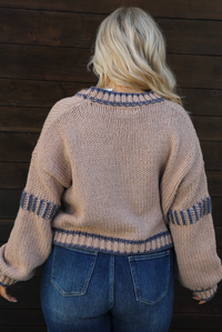 Woman model wearing a mocha brown cardigan that has grey stitching details throughout. The cardigan has a cropped cut, is oversized, and has functional button closures in the front. She's wearing the cardigan with dark denim jeans. The photo showcases a close up of the back of the cardigan. The photo is taken against a dark brown wooden door outdoors. 