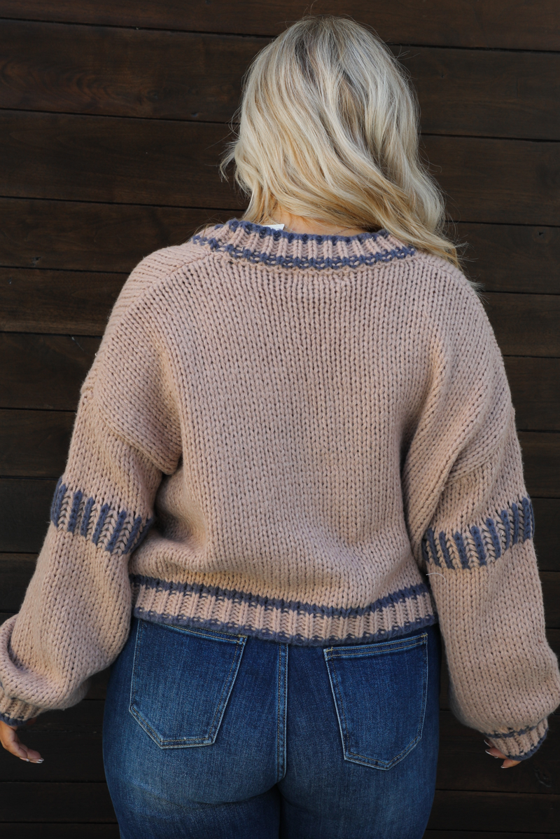 Woman model wearing a mocha brown cardigan that has grey stitching details throughout. The cardigan has a cropped cut, is oversized, and has functional button closures in the front. She's wearing the cardigan with dark denim jeans. The photo showcases a close up of the back of the cardigan. The photo is taken against a dark brown wooden door outdoors. 