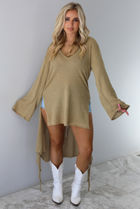 Woman wearing a camel long-sleeve top that's tied in the front with a knot and has a train in the back. The top is untied to show the full length of the front, which stops mid-thigh on the 5'8" model. The photo is taken indoors against a white wall with white marble flooring. 
