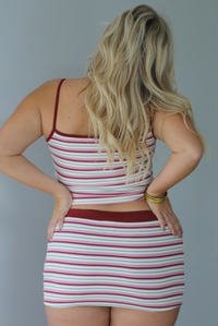 A woman shown from behind wearing a berry, pink, white, and grey striped two piece set featuring a tank with a mini skirt, standing indoors against a white wall. 