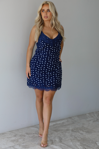 Woman wearing a navy dress with a white polka dot print pattern throughout with blue lace detailing at the neckline and hem indoors against a white wall with white marble flooring. 
